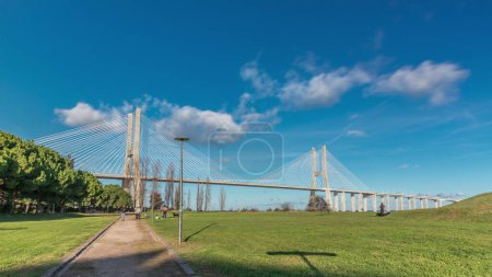 Téléchargez les photos : The Vasco da Gama Bridge timelapse hyperlapse with green lawn and trees. Cable-stayed longest bridge flanked by viaducts and rangeviews that spans Tagus River in Park of Nations in Lisbon, Portugal - en image libre de droit
