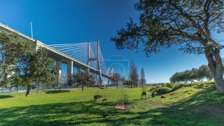 Téléchargez les photos : The Vasco da Gama Bridge timelapse hyperlapse viewed from green lawn with trees. Cable-stayed longest bridge flanked by viaducts and rangeviews that spans Tagus River in Park Nations. Lisbon, Portugal - en image libre de droit