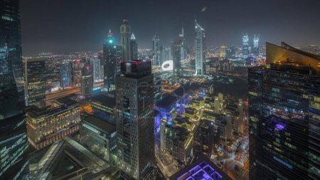 Foto de Panorama mostrando rascacielos futuristas en el centro de negocios del distrito financiero en Dubai en Sheikh Zayed carretera noche timelapse. Vista aérea desde arriba con torres iluminadas - Imagen libre de derechos