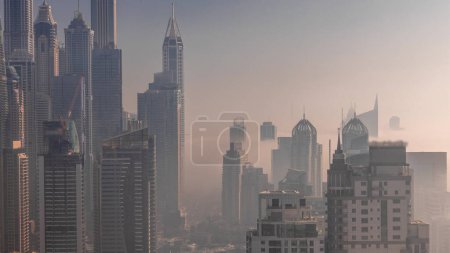 Foto de Vista de varios rascacielos en el bloque residencial más alto de Dubai Marina durante el amanecer timelapse aéreo con canal artificial. Niebla matutina entre muchas torres y yates - Imagen libre de derechos
