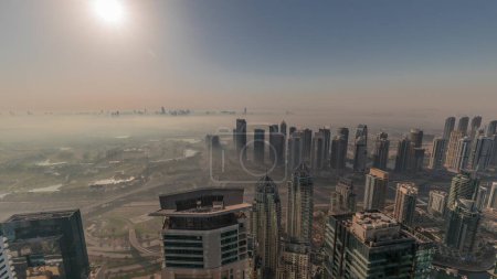 Téléchargez les photos : Panorama de Dubai Marina avec gratte-ciel JLT et parcours de golf pendant le lever du soleil timelapse, Dubaï, Émirats arabes unis. Vue aérienne depuis les tours matin brumeux. Skyline de la ville avec ciel orange - en image libre de droit