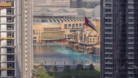 Téléchargez les photos : Vue aérienne de la fontaine de Dubaï au centre-ville avec palmiers et drapeau dans le parc à côté du centre commercial et souq timelapse, Émirats Arabes Unis. Vu entre deux gratte-ciel d'en haut. Ombres longues se déplaçant rapidement - en image libre de droit