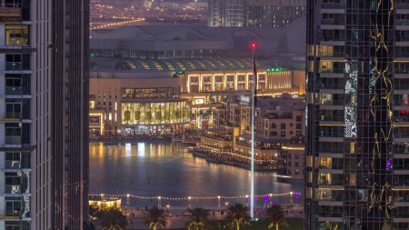 Téléchargez les photos : Vue aérienne de la fontaine de Dubaï au centre-ville avec des palmiers et un drapeau dans le parc à côté du centre commercial et du souk Timelapse de transition du jour à la nuit, Émirats arabes unis. Vu entre deux gratte-ciel d'en haut après le coucher du soleil - en image libre de droit