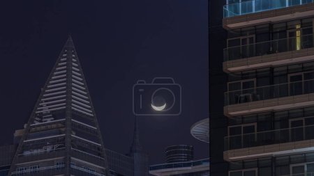 Foto de Crescent Moon está detrás de rascacielos en Dubai timelapse. Majestuosa vista durante la noche en la ciudad. Vista aérea de torres superiores con luz en ventanas. Distrito de Business Bay - Imagen libre de derechos