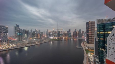 Foto de Skyline aéreo de Dubai Business Bay y el centro de la ciudad con los diversos rascacielos y torres a lo largo de la costa en el canal de día a la noche timelapse transición. Lugar de construcción con grúas después del atardecer - Imagen libre de derechos