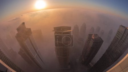 Téléchargez les photos : Brouillard hivernal rare tôt le matin au lever du soleil au-dessus de la ligne d'horizon de Dubai Marina et JLT et gratte-ciel éclairés par des lampadaires transition aérienne de nuit au jour. Vue de dessus depuis les nuages. Dubai, Émirats arabes unis - en image libre de droit