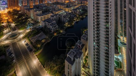 Téléchargez les photos : Bâtiments d'étang et de faible hauteur dans l'antenne du quartier des Verts pendant toute la nuit. Skyline de Dubaï avec des gratte-ciel dans le quartier de Barsha Heights sur un fond - en image libre de droit