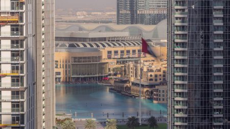 Foto de Vista aérea de la Fuente de Dubái en el centro con palmeras y bandera en el parque junto al centro comercial y el timelapse zoco, Emiratos Árabes Unidos. Visto entre dos rascacielos de arriba. Largas sombras moviéndose rápido - Imagen libre de derechos