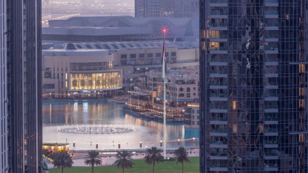 Foto de Vista aérea de Dubai Fuente en el centro con palmeras y bandera en el parque junto al centro comercial y zoco noche a día timelapse transición, Emiratos Árabes Unidos. Visto entre dos rascacielos desde arriba antes del amanecer - Imagen libre de derechos