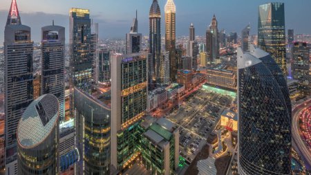 Foto de Vista panorámica de los edificios de gran altura en Sheikh Zayed Road en Dubái día aéreo a noche timelapse transición, Emiratos Árabes Unidos. Rascacielos iluminados y estacionamiento en el Centro Financiero Internacional desde arriba - Imagen libre de derechos