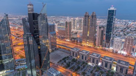 Foto de Edificios de gran altura en Sheikh Zayed Road en Dubái noche aérea al día timelapse transición, Emiratos Árabes Unidos. Rascacielos en el distrito financiero internacional desde arriba. Paseo por la ciudad casas y villas en el fondo - Imagen libre de derechos