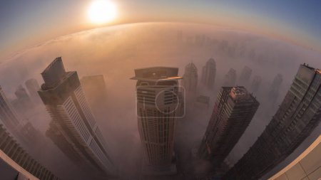 Téléchargez les photos : Brouillard hivernal rare tôt le matin au lever du soleil au-dessus de la ligne d'horizon de Dubai Marina et JLT et gratte-ciel éclairés par des lampadaires transition aérienne de nuit au jour. Vue de dessus depuis les nuages. Dubai, Émirats arabes unis - en image libre de droit