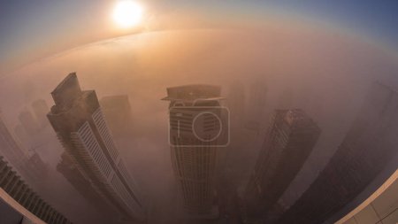 Foto de Rara niebla invernal temprano en la mañana durante el amanecer por encima del horizonte de Dubai Marina y JLT y rascacielos iluminados por luces de la calle noche aérea a la transición del día. Vista superior desde las nubes superiores. Dubai, Emiratos Árabes Unidos - Imagen libre de derechos