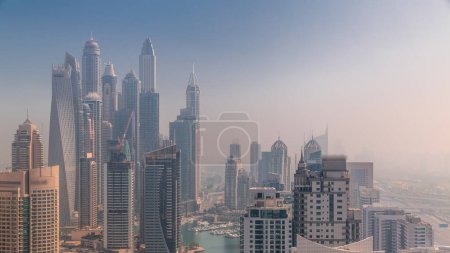 Foto de Vista de varios rascacielos en el bloque residencial más alto de Dubai Marina durante el amanecer timelapse aéreo con canal artificial. Muchas torres y yates cubiertos por la niebla de la mañana - Imagen libre de derechos