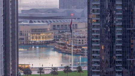Téléchargez les photos : Vue aérienne de la fontaine de Dubaï au centre-ville avec palmiers et drapeau dans le parc à côté du centre commercial et souk nuit à jour timelapse transition, Émirats Arabes Unis. Vu entre deux gratte-ciel d'en haut avant le lever du soleil - en image libre de droit