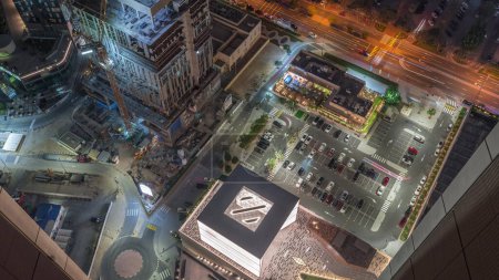 Foto de Sitio de construcción con grúas y estacionamiento cerca de la avenida comercial en Dubai timelapse noche aérea, Emiratos Árabes Unidos. Torres en el distrito financiero desde arriba. Tráfico en una carretera - Imagen libre de derechos