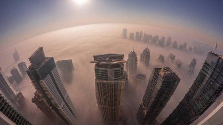 Téléchargez les photos : Brouillard hivernal rare tôt le matin au lever du soleil au-dessus des toits de Dubai Marina et JLT et des gratte-ciel éclairés par des lampadaires aériens. Vue de dessus depuis les nuages. Dubai, Émirats arabes unis - en image libre de droit