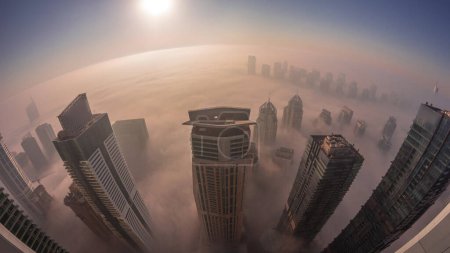 Photo for Rare early morning winter fog during sunrise above the Dubai Marina and JLT skyline and skyscrapers lighted by street lights aerial. Top view from above clouds. Dubai, UAE - Royalty Free Image