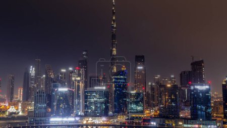 Téléchargez les photos : Vue aérienne vers Dubai Business Bay et le centre-ville toute la nuit avec les différents gratte-ciel et tours le long du front de mer sur le canal timelapse de nuit. Site de construction avec grues. Nuages et brouillard - en image libre de droit