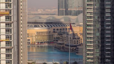 Foto de Vista aérea de la Fuente de Dubái en el centro con palmeras y bandera en el parque junto al centro comercial y el timelapse zoco, Emiratos Árabes Unidos. Visto entre dos rascacielos de arriba. Largas sombras moviéndose rápido - Imagen libre de derechos