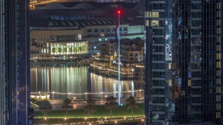 Foto de Vista aérea de la Fuente de Dubái en el centro con palmeras y bandera en el parque al lado del centro comercial y timelapse zoco durante toda la noche, Emiratos Árabes Unidos. Visto entre dos rascacielos desde arriba con las luces apagadas - Imagen libre de derechos