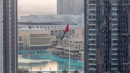 Téléchargez les photos : Vue aérienne de la fontaine de Dubaï au centre-ville avec palmiers et drapeau dans le parc à côté du centre commercial et souk nuit à jour timelapse transition, Émirats Arabes Unis. Vu entre deux gratte-ciel d'en haut avant le lever du soleil - en image libre de droit
