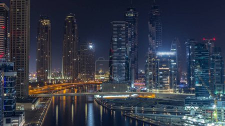 Foto de Paisaje urbano con rascacielos iluminados de Dubai Business Bay y antena de canal de agua durante toda la noche. skyline moderno con torres residenciales y de oficina en el paseo marítimo. Luna poniéndose - Imagen libre de derechos