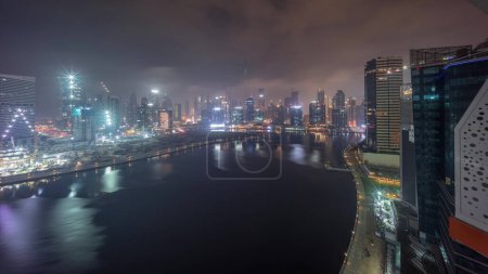Téléchargez les photos : Vue aérienne vers Dubai Business Bay et le centre-ville avec les différents gratte-ciel et tours couverts de brouillard toute la nuit. Timelapse nocturne au bord de l'eau et du canal. Site de construction avec grues - en image libre de droit