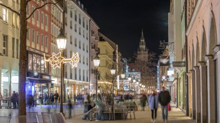 Téléchargez les photos : Bâtiments historiques illuminés dans la vieille ville de Munich - sendlinger strasse night timelapse. Rue piétonne avec bancs et lampadaires. Glockenspiel sur un fond. Allemagne - en image libre de droit
