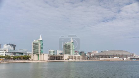 Téléchargez les photos : Modern tall residential and office buildings reflecting in the water under blue cloudy sky timelapse hyperlapse in Lisbon, Portugal. Park nations luxury district with concert hall and flags - en image libre de droit