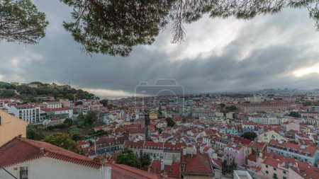 Panorama showing aerial cityscape day to night transition from Miradouro da Graca viewpoint in Lisbon city after sunset. Dramatic clouds over historic houses with red roofs and evening illumination