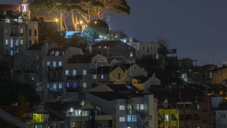 Téléchargez les photos : Point de vue célèbre Miradouro da Senhora do Monte avec église et vue sur la ville de Lisbonne timelapse avec des lumières dans les fenêtres de maisons colorées autour, Lisbonne, Portugal. Arbres verts sur le dessus - en image libre de droit