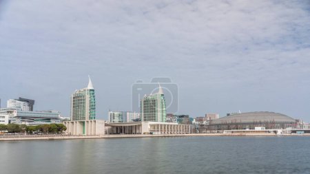 Photo for Modern tall residential and office buildings reflecting in the water under blue cloudy sky timelapse hyperlapse in Lisbon, Portugal. Park nations luxury district with concert hall and flags - Royalty Free Image