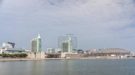 Téléchargez les photos : Modern tall residential and office buildings reflecting in the water under blue cloudy sky timelapse hyperlapse in Lisbon, Portugal. Park nations luxury district with concert hall and flags - en image libre de droit