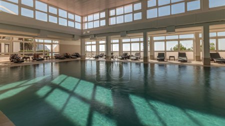 Foto de Panorama que muestra la lujosa piscina cubierta, parte de la zona de spa del hotel timelapse. Luz solar a través de ventanas panorámicas con océano y nubes en un cielo azul - Imagen libre de derechos