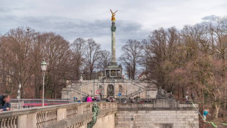 Foto de Columna de la Paz con estatua de Ángel de la Paz de oro (Friedensengel) timelapse, la gente en el puente sobre el río Isar mirando al caminante de cuerda. Parque público en la capital bávara. Alemania, Múnich, Bogenhausen - Imagen libre de derechos
