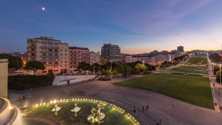 Panorama montrant la pelouse à Alameda Dom Afonso Henriques avec des bâtiments colorés et illuminés Fontaine lumineuse transition aérienne jour-nuit timelapse d'en haut après le coucher du soleil à Lisbonne, Portugal