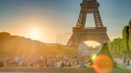 Téléchargez les photos : Tour Eiffel à partir de Champ de Mars pendant le coucher du soleil timelapse, Paris, France. Des gens assis sur la pelouse. Soirée d'été avec ciel dégagé. Ciel orange et rayons de lumière - en image libre de droit