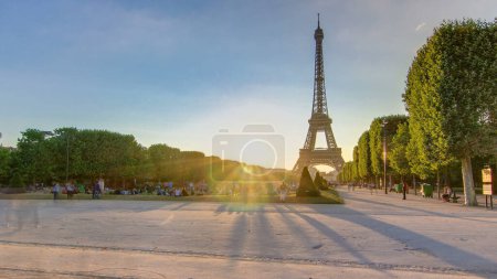 Photo for Eiffel Tower seen from Champ de Mars during sunset timelapse, Paris, France. People sitting on lawn and walking around. Summer evening with clear sky and sun beams - Royalty Free Image