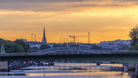 Eisenbahnbrücke (Pont Rouelle) über Insel (Ile des Cygnes) Zeitraffer am Morgen nach Sonnenaufgang in Paris. Orangefarbener Himmel und Spiegelung auf dem Wasser der Seine. Baukräne