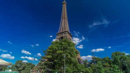 Photo for Eiffel Tower close up view from Siene river waterfront in Paris timelapse hyperlapse, France. Blue cloudy sky at summer day with green trees and people walking around - Royalty Free Image