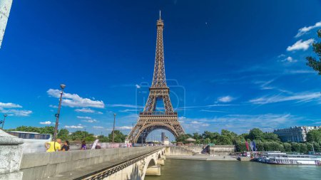 Foto de Torre Eiffel con puente sobre el río Siene en Paris timelapse hyperlapse, Francia. Cielo azul nublado en el día de verano con árboles verdes y gente caminando sobre el puente - Imagen libre de derechos