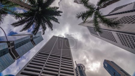 Foto de Mirando hacia arriba la perspectiva de los rascacielos de negocios modernos de vidrio y vista al cielo paisaje de edificio comercial en el timelapse de la ciudad central. Torres con reflejos y palmeras en Singapur - Imagen libre de derechos