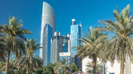 Téléchargez les photos : Corniche boulevard plage parc le long de la côte à Abu Dhabi timelapse avec des gratte-ciel en arrière-plan. Vue depuis le parc familial. Les paumes sur le côté. Ciel bleu au soleil - en image libre de droit
