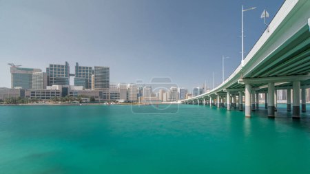 Foto de Edificios modernos en Abu Dhabi skyline timelapse hiperlapso con centro comercial y playa. Vista desde el paseo marítimo de la isla Al Maryah con reflejos en el agua cerca del puente - Imagen libre de derechos