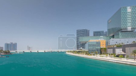 Photo for Modern buildings of Al Maryah Island in Abu Dhabi skyline timelapse hyperlapse with mall and promenade. View from the bridge with reflections of skyscrapers in water - Royalty Free Image