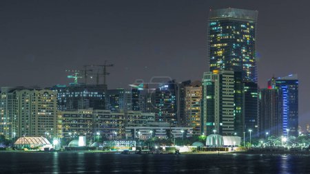 Foto de Vista panorámica del horizonte de Abu Dhabi y frente al mar por la noche timelapse, Emiratos Árabes Unidos. Vista de Corniche con rascacielos iluminados y obras de construcción - Imagen libre de derechos
