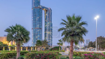 Foto de Rascacielos de Abu Dhabi iluminados con torres Nation edificios día a noche timelapse transición. Palmeras en parque y farola - Imagen libre de derechos