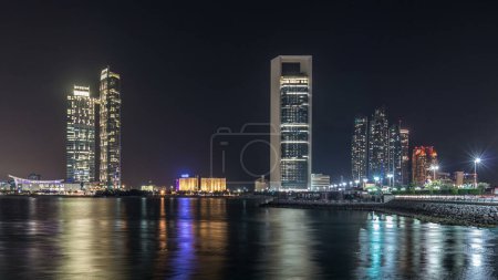 Foto de Vista panorámica del horizonte de Abu Dhabi y frente al mar por la noche timelapse, Emiratos Árabes Unidos. Vista de Corniche con rascacielos iluminados reflejados en el agua - Imagen libre de derechos