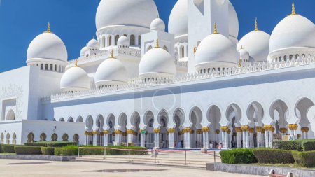 Téléchargez les photos : Timelapse de la Grande Mosquée Cheikh Zayed à Abu Dhabi, la capitale des Émirats arabes unis. Vue latérale avec fontaine, dômes et minarets. Ciel bleu au soleil - en image libre de droit